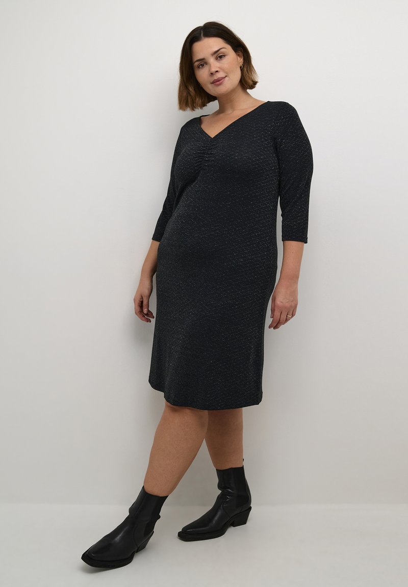 Robe noire texturée, longueur genou, avec un décolleté en V et des manches trois-quarts. Portée avec des bottines noires, se tenant contre un fond blanc.