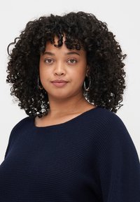 Le pull en tricot bleu marine présente un col large et un motif texturé. Le mannequin a les cheveux bouclés et porte des boucles d'oreilles créoles.