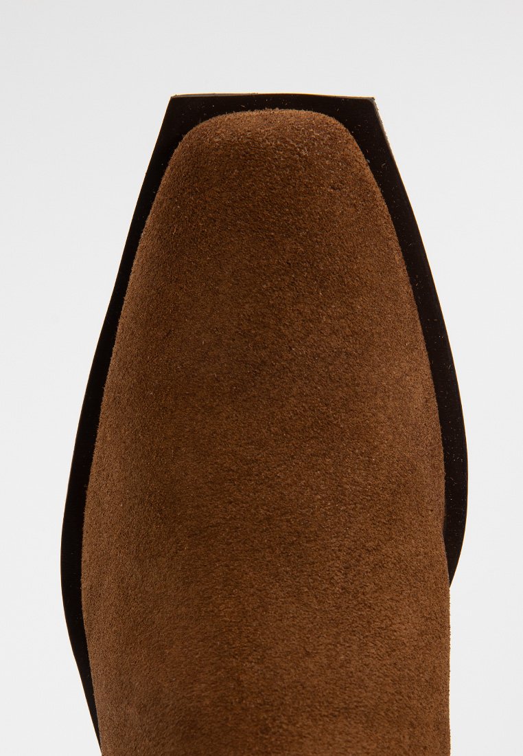 Bruine suède laarzenpunt met een vierkante zwarte zoolrand, gezien vanuit een top-down perspectief op een witte achtergrond.