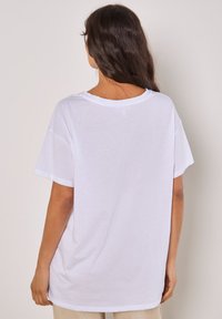 Weißes Baumwoll-T-Shirt mit lockerer Passform, kurzen Ärmeln und weitem Ausschnitt. Glatte Textur mit minimalen Nahtdetails am Saum.