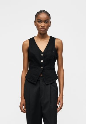 Giovane donna che indossa un gilet nero senza maniche con bottoni e tasca e pantaloni neri plissettati, in piedi davanti a uno sfondo chiaro uniforme.