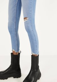 Jeans skinny azul claro con desgastes en las rodillas y dobladillos deshilachados, combinados con botas negras hasta el tobillo con suela gruesa y lados elásticos.