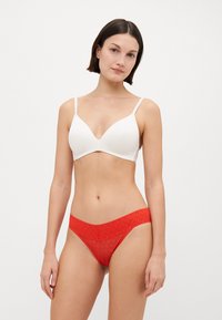 Soutien-gorge blanc rembourré avec des fines bretelles, présentant un décolleté plongeant. String rouge avec une large taille décorative et une texture à motifs.