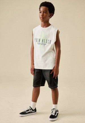 Chlapec v bielom tielku s potlačou "Palm Beach", čiernych krátkych nohaviciach, bielych ponožkách a čiernych teniskách, stojaci pred jednofarebným béžovým pozadím.
