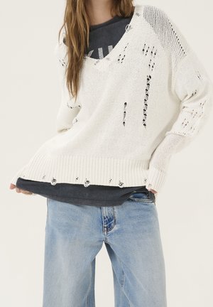 Personne portant un pull en maille blanc ample avec des trous intentionnels sur un t-shirt noir et un jean bleu clair, debout devant un fond uni.