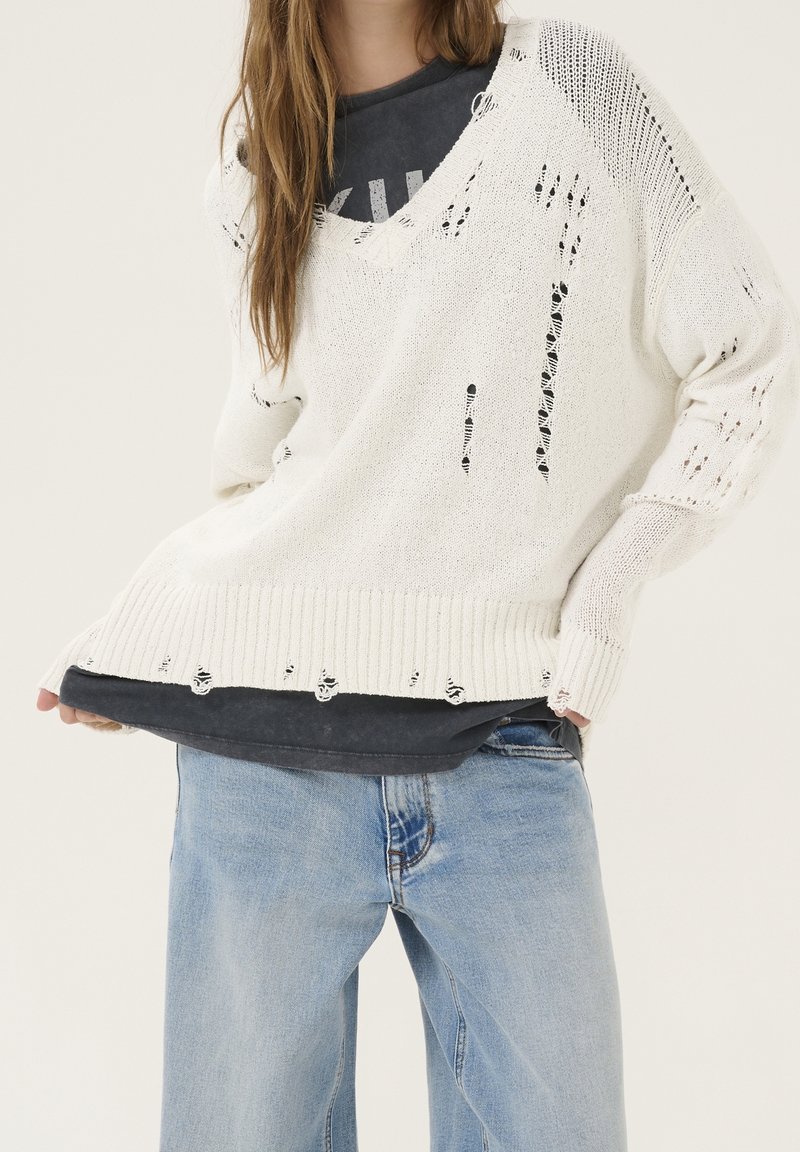 Personne portant un pull en maille blanc ample avec des trous intentionnels sur un t-shirt noir et un jean bleu clair, debout devant un fond uni.