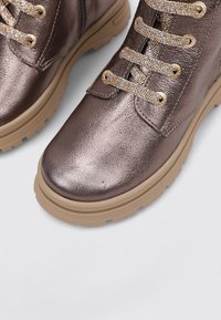 Bottes chevilles en bronze métallisé avec une surface texturée, semelle en caoutchouc beige et lacets brillants et ornés. Design à bout rond.