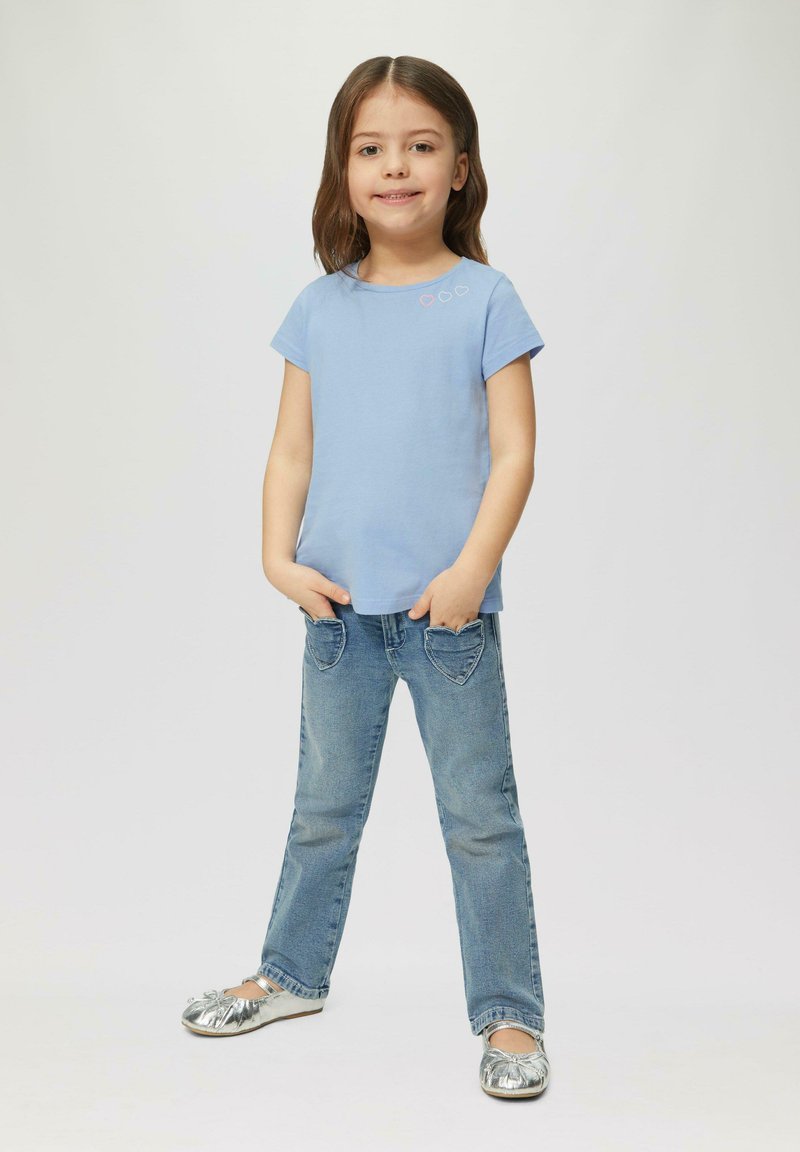 Helles blaues Baumwoll-T-Shirt mit kurzen Ärmeln und drei kleinen Herzverzierungen. Getragen mit blauen Jeans mit herzförmigen Taschen und silbernen Schuhen.