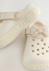 Sabots couleur crème avec une finition lisse, présentant des côtés fendus, un accent en nœud et le logo "BAKER" sur la semelle et la bride.
