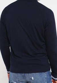 Felpa girocollo marina con polsini e fondo a costine, caratterizzata da una texture liscia e maniche raglan lunghe, indossata sopra jeans strappati.