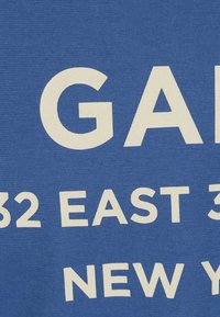 Blauwe katoenen stof met grote witte tekst. Tekst omvat "GAP," "32 EAST 32," en "NEW YORK." Eenvoudig ontwerp, gladde textuur.