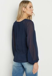 Mörkblå blus med genomskinliga långa ärmar och texturerat prickmönster. Nyckelhålsknäppning bak och lös passform. Matchas med ljus denim.