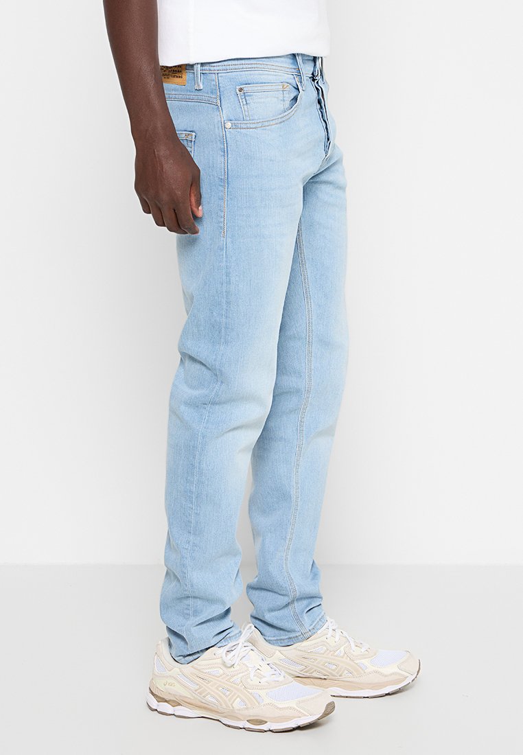 Persoon in lichtblauwe jeans en witte sneakers die tegen een effen witte achtergrond staat, zijaanzicht.