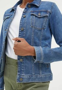 Person wearing blue denim jacket over white shirt and olive green pants, with hand holding jacket near chest.