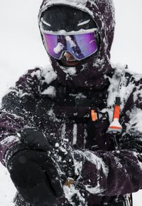 Person in purple winter gear and purple goggles covered in snow, holding snow with gloved hands in a snowy outdoor setting.