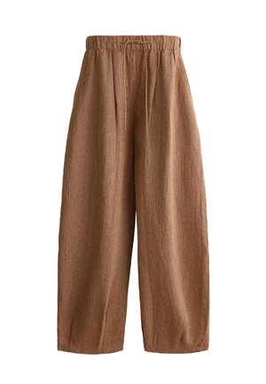Młoda kobieta z falującymi włosami, ubrana w biały bezrękawnik typu crop top, brązowe szerokie spodnie, związaną bluzę wokół talii i klapki, idzie po kafelkowanej podłodze.