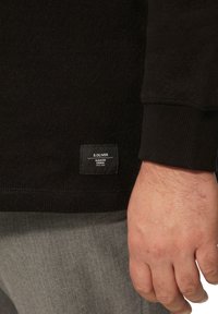 Zwarte lange mouwen shirt met ribbeltextuur, met een zichtbaar S. Oliver label op de zoom en grijze gebloemde broek. Hand in het frame.