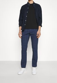 Camisa azul marino abotonada, camiseta negra y pantalones de cuadros azul marino. Zapatillas blancas completan el atuendo, mostrando un estilo casual moderno.