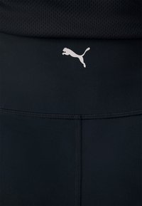 Musthaidistants liigpüksid, millel on sile ja veniv kangas ning valge Puma logo, mis on paigutatud vöökohtadele.