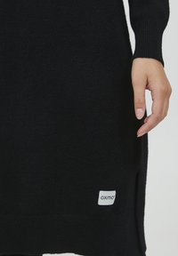 Schwarzer Pullover mit gerippten Bündchen und seitlichen Schlitzen. Mit einem kleinen weißen Etikett mit "oxmo" am unteren Saum. Weicher, strukturiert Stoff.