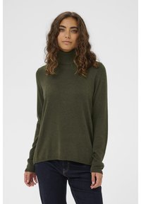 Tumši zaļš augstu apkaklītis džemperis ar garām piedurknēm, izgatavots no mīksta materiāla. Raksturīga vaļīga piegriezuma un ribotas aproces, kopā ar tumšām džinsa biksēm.