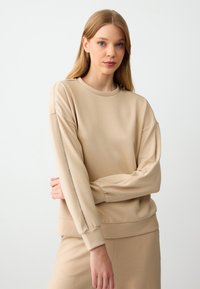 Felpa beige a maniche lunghe con scollatura rotonda e una consistenza morbida. Presenta dettagli metallici lungo le maniche e una vestibilità comoda.
