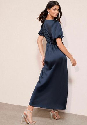 Navy-satin maxi-mekko, jossa on puff-hihat, vyötäröltä kiristetty malli, sileä pinta ja pitkä siluetti, yhdistettynä hopeisiin korkokenkiin.