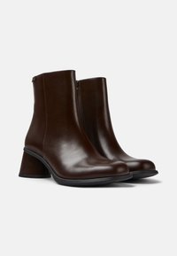 Paire de bottines en cuir marron foncé à talons carrés et fermetures éclair latérales, présentées sur un fond clair uni.
