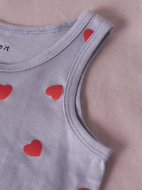 Nahaufnahme eines hellvioletten ärmellosen Tops mit roten Herzmustern, auf einem hellrosa Stoffhintergrund gelegt.