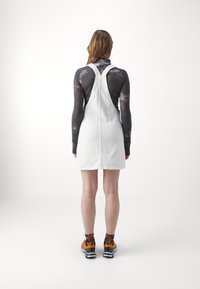 Biała sukienka ogrodniczka z denimu z regulowanymi szelkami, kieszeniami na biodrach i prostym krojem, zestawiona z kolorowymi skarpetkami w wzory i pomarańczowymi trampkami.