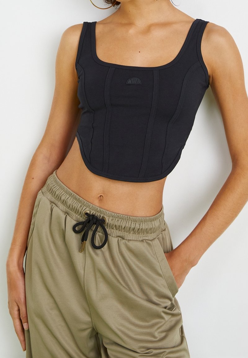 Femme portant un crop top noir sans manches et un pantalon ample kaki avec un cordon noir, main dans la poche, sur fond clair uni.