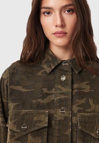 Junge Frau, die eine olivgrüne Cargomuster-Jacke aus Kordsamt mit Metallknöpfen und Fronttaschen trägt, die mit dem Gesicht zur Kamera vor einem einfarbigen Hintergrund steht.
