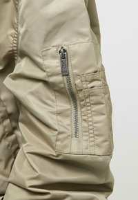 Närbild av en beige jackärm med en vertikal dragkedjeficka och sydda detaljer på tyget.