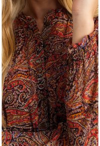 Femme portant une chemise boutonnée à manches longues avec un motif cachemire complexe rouge, orange, noir et blanc et un col à volants.