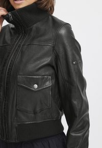 Černá kožená bunda s žebrovaným límcem, dvěma předními kapsami, stříbrným drukem a přiléhavým střihem. Hladká textura a strukturovaný silueta.