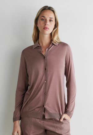 Frau mit langen blonden Haaren trägt eine mauvefarbene, geknöpfte Strickjacke und passende Hose, steht vor einer weißen Wand mit einer Hand in der Tasche.