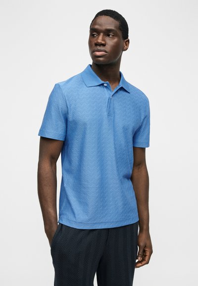 Μαύρος άνδρας που φοράει ανάγλυφο ανοιχτό μπλε πόλο με κοντό μανίκι και σκουρόχρωμο υφασμάτινο παντελόνι, στέκεται με τα χέρια στις τσέπες, κοιτάζοντας προς τα πλάγια.