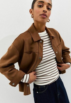 Kobieta ubrana w brązową krótką kurtkę na białej i brązowej paskowanej bluzce z długim rękawem, niebieskie dżinsy i kolczyki-koła na białym tle.