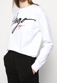 Felpa bianca con logo nero e rosso, maniche lunghe con polsini a costine, taglio rilassato, realizzata in tessuto morbido, design minimalista.