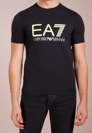 Uomo che indossa una t-shirt nera a maniche corte con logo "EA7" giallo neon e "Emporio Armani" bianco sul petto, abbinata a pantaloni neri.
