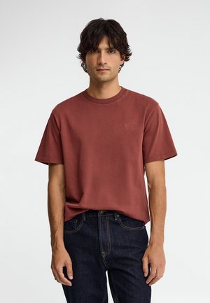 Uomo con capelli scuri che indossa una maglietta tinta unita color ruggine e jeans blu scuro, in piedi davanti a uno sfondo bianco uniforme.