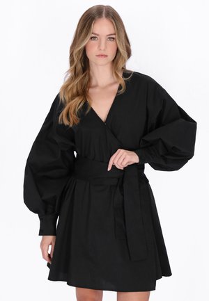 Giovane donna con capelli lunghi e mossi che indossa un vestito nero con scollo a V, maniche a palloncino oversize e cintura in vita, in piedi davanti a uno sfondo bianco.