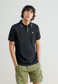 Sort polo t-shirt med hvid kravekant og et lille logo, parret med grønne cargo shorts med sidelommer og en afslappet pasform.