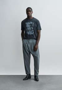 Junger Mann steht mit den Händen in den Taschen, trägt ein schwarzes T-Shirt mit Grafik, graue plissierte Hosen und schwarze Knöchelstiefel auf einem grauen Boden.