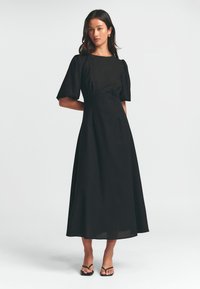 Černé šaty střední délky s krátkými nabíranými rukávy, kulatým výstřihem, přiléhavým korzetem a splývavou sukni. Oblečené s jednoduchými černými sandály.