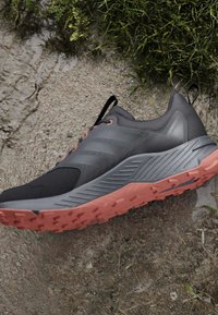 Chaussure de trail running gris foncé et noire avec semelle corail, posée sur un sol boueux à côté d’herbe verte.