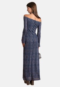 Langes, schulterfreies Kleid in Marineblau mit aufwendigen Blumenmustern, langen Ärmeln und taillierter Silhouette, kombiniert mit Absatzstiefeletten.