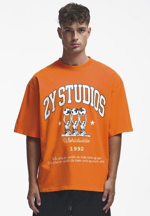 Giovane uomo che indossa una maglietta oversize arancione brillante con la scritta "ZY STUDIOS Worldwide 1992" e la grafica di tre figure che tengono dei globi.