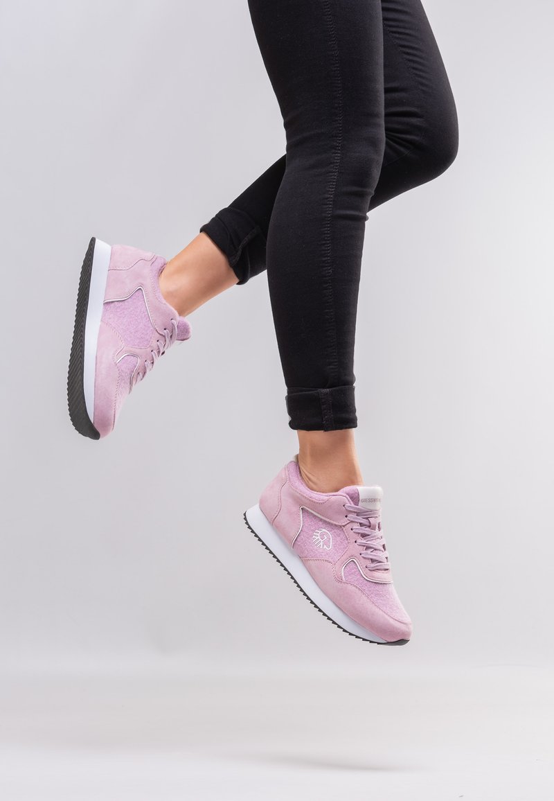 Persoon in een zwarte broek springt en toont pastelroze sneakers met witte zolen tegen een effen lichtgrijze achtergrond.