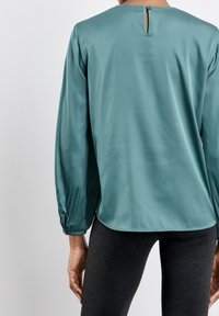 Blusa di raso verde acqua con scollatura a girocollo, maniche lunghe a palloncino con polsini elasticizzati e una piccola apertura a oblò sul retro.